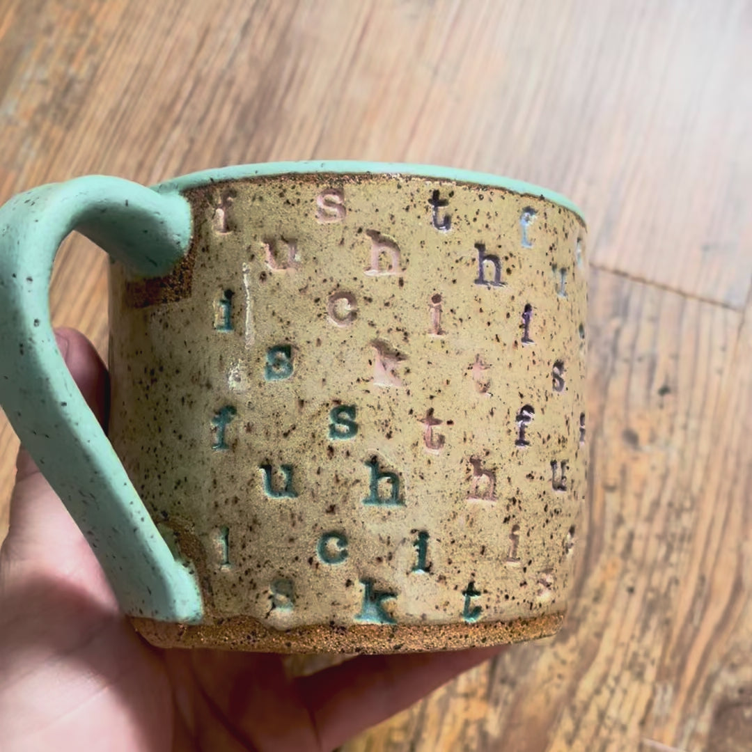 A video rotating around a mug stamped with "f*ck this sh*t" in diagonal stripes with a seaglass colored handle