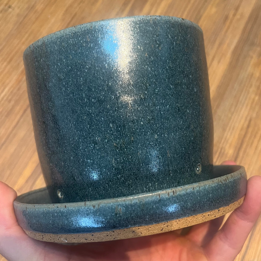 A handmade ceramic planter with a wide saucer base, held in a hand against a wooden floor background. The planter has a speckled dark teal-blue glaze with a natural beige clay showing at the rim and base edge.