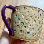 A handmade ceramic mug with a purple handle and rim, featuring a speckled beige body with embossed letters and stars in blue and pink, held by a hand over a wooden surface.