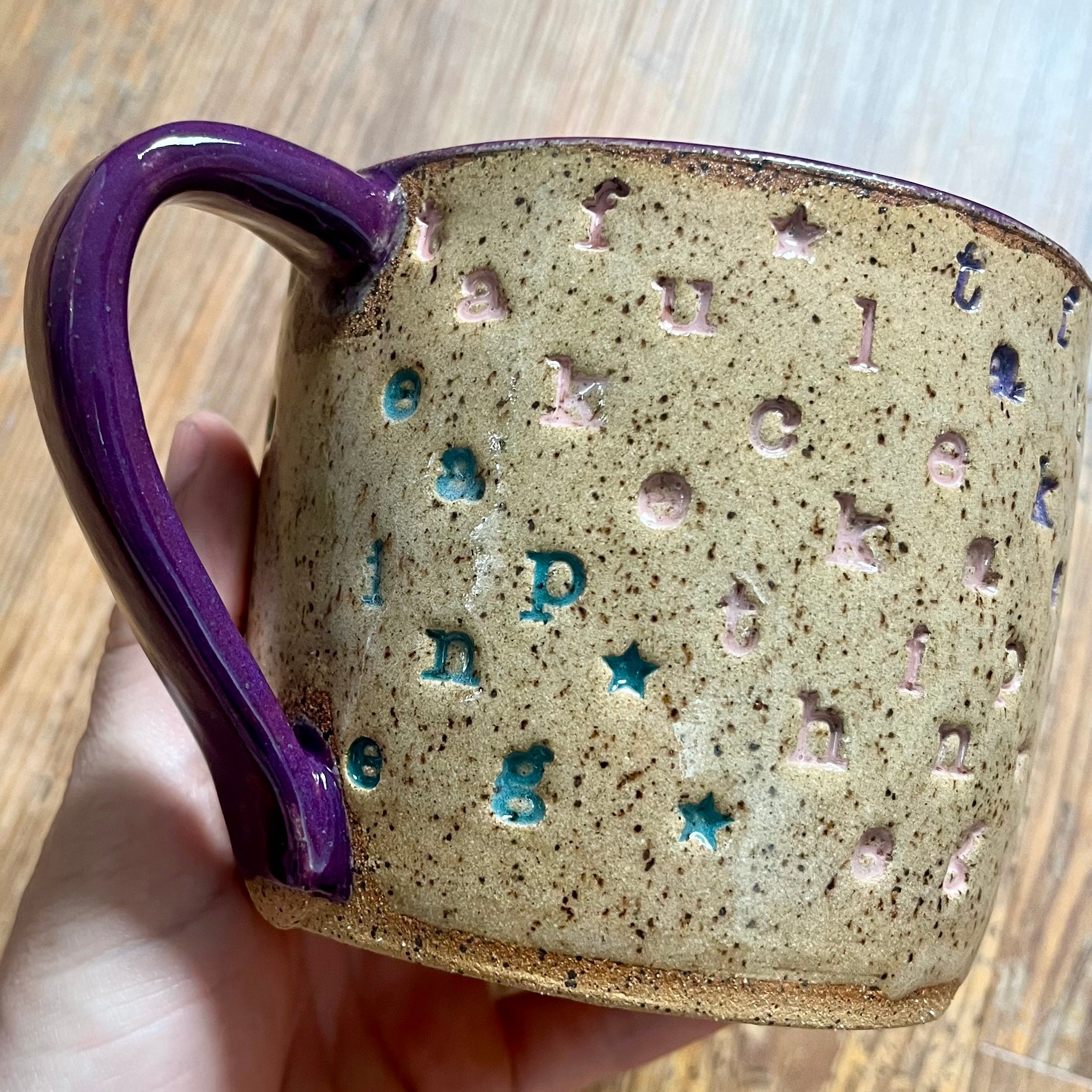 A handmade ceramic mug with a purple handle and rim, featuring a speckled beige body with embossed letters and stars in blue and pink, held by a hand over a wooden surface.
