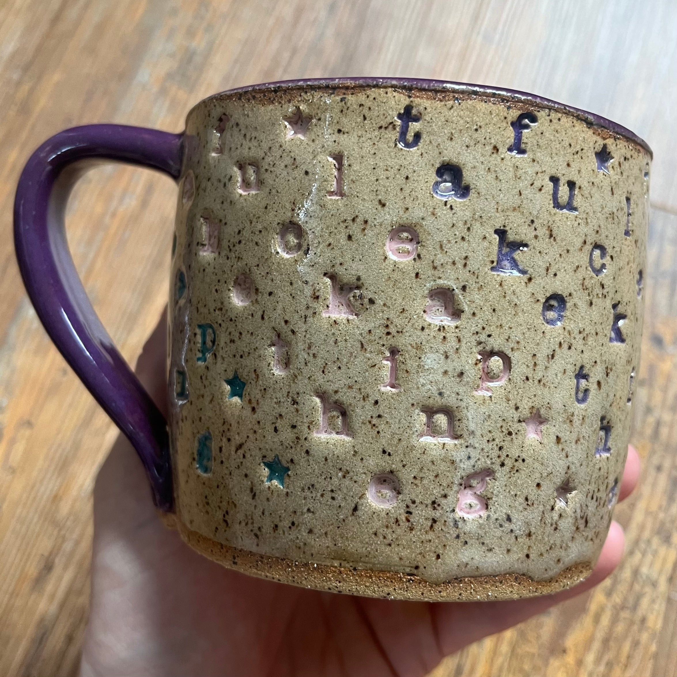 A handmade ceramic mug with a textured beige and brown speckled glaze, featuring embossed letters spelling out words and small star cutouts. The mug has a glossy purple handle and rim, and is being held by a hand over a wooden table background.