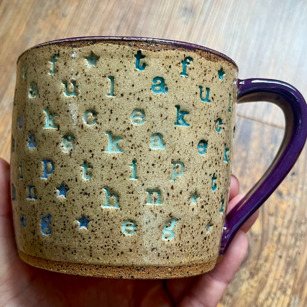 A handmade ceramic mug with a speckled beige and brown glaze, featuring embossed turquoise blue letters and stars, held in a hand over a wooden table background.