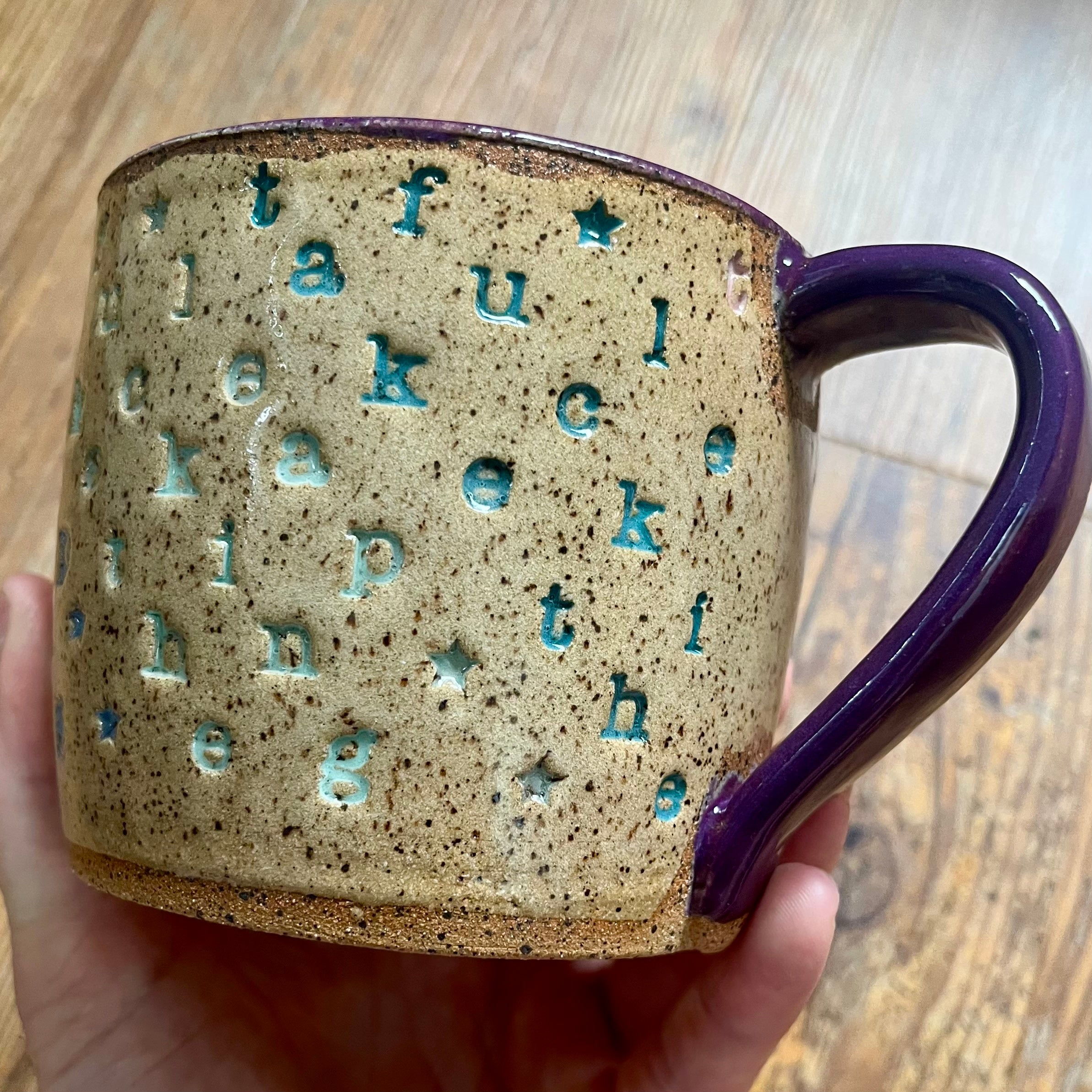 A handmade ceramic mug with a speckled beige and brown glaze, featuring raised letters spelling out 'thankful' and small star motifs in turquoise, held in a hand against a wooden background.