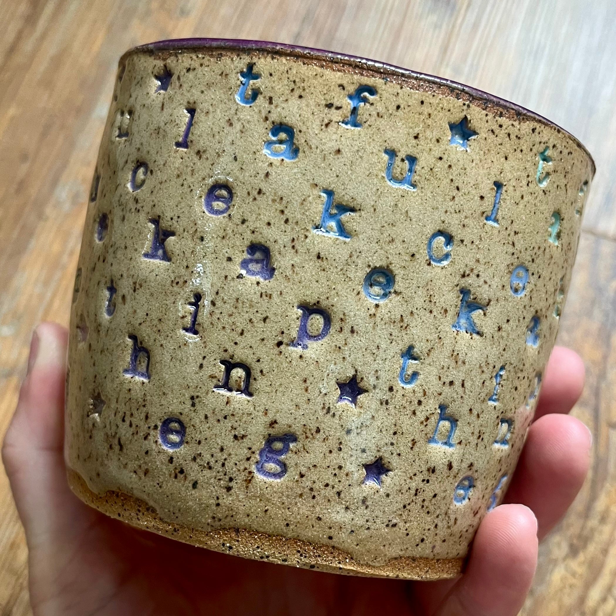 A handmade ceramic cup held in a hand, featuring a speckled beige glaze with cutout letters spelling out words and small blue star accents, set against a wooden background.