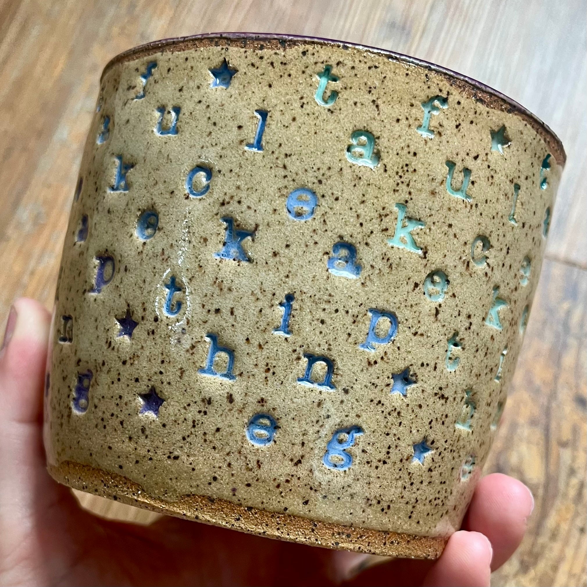 A handmade ceramic cup held in a hand, featuring a speckled beige-brown glaze with embossed blue letters and stars, set against a wooden background.