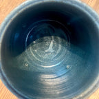 An overhead close-up view of the inside of a dark ceramic planter, showing a speckled blue-gray glaze, small circular marks on the bottom, and a repaired crack, set against a wooden surface.