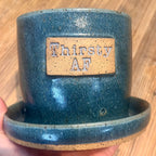 A handmade ceramic planter in a speckled teal-blue glaze with an attached saucer, held by a hand against a wooden floor background. The planter features a tan speckled plate on the front with the words 'Thirsty AF' embossed.