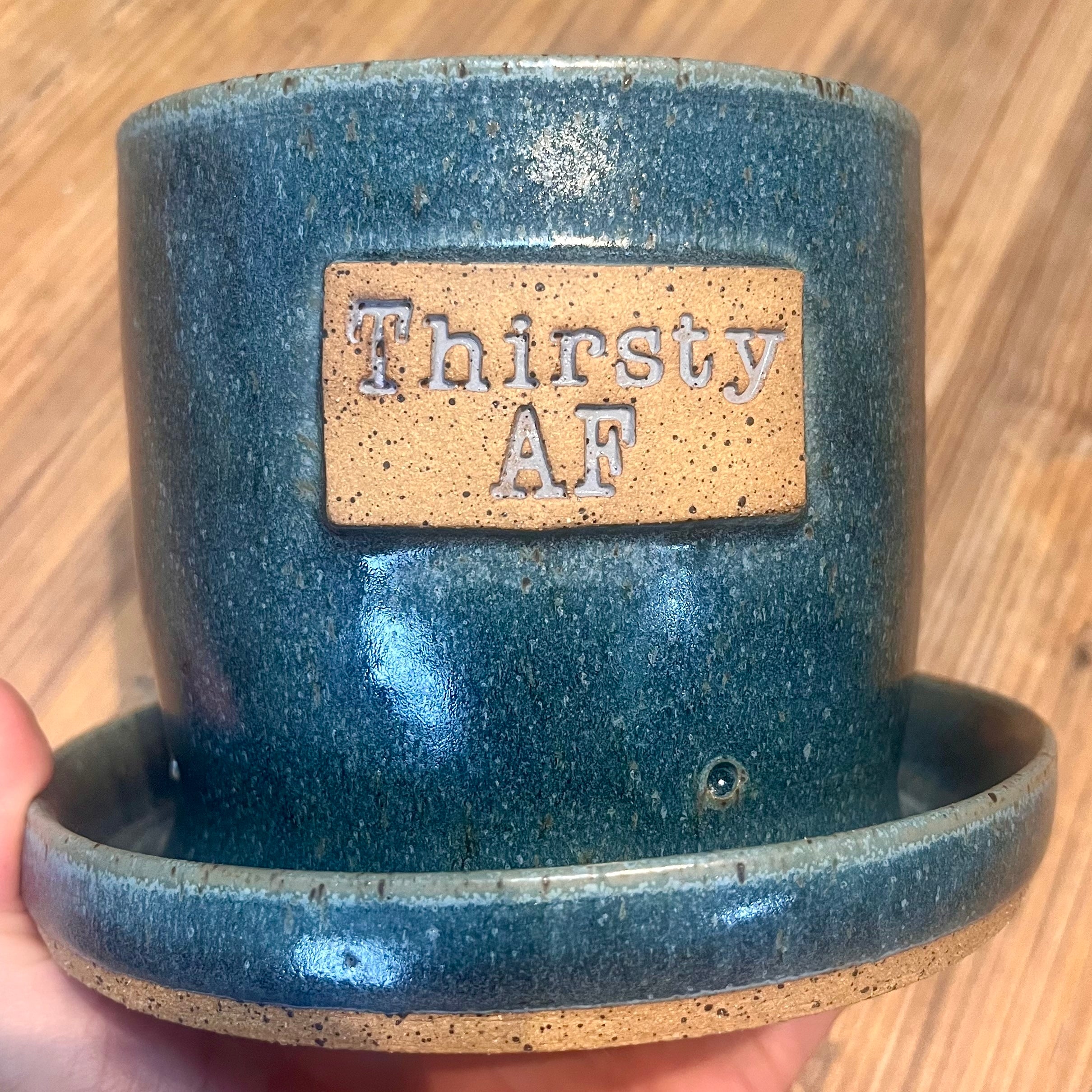 A handmade ceramic planter in a speckled teal-blue glaze with an attached saucer, held by a hand against a wooden floor background. The planter features a tan speckled plate on the front with the words 'Thirsty AF' embossed.