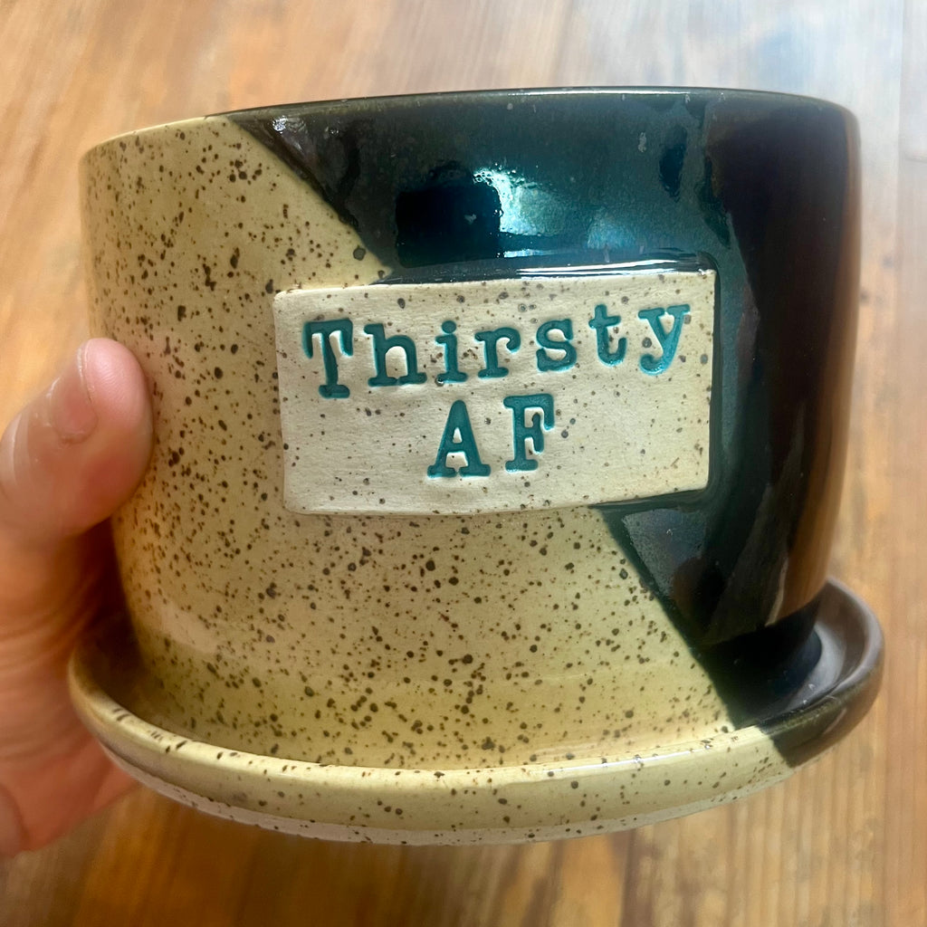 A ceramic planter pot with an attached saucer, held by a hand. The pot features a cream-colored speckled glaze on the left and a glossy black glaze on the right, with a rectangular cream label on the front that reads 'Thirsty AF' in green.