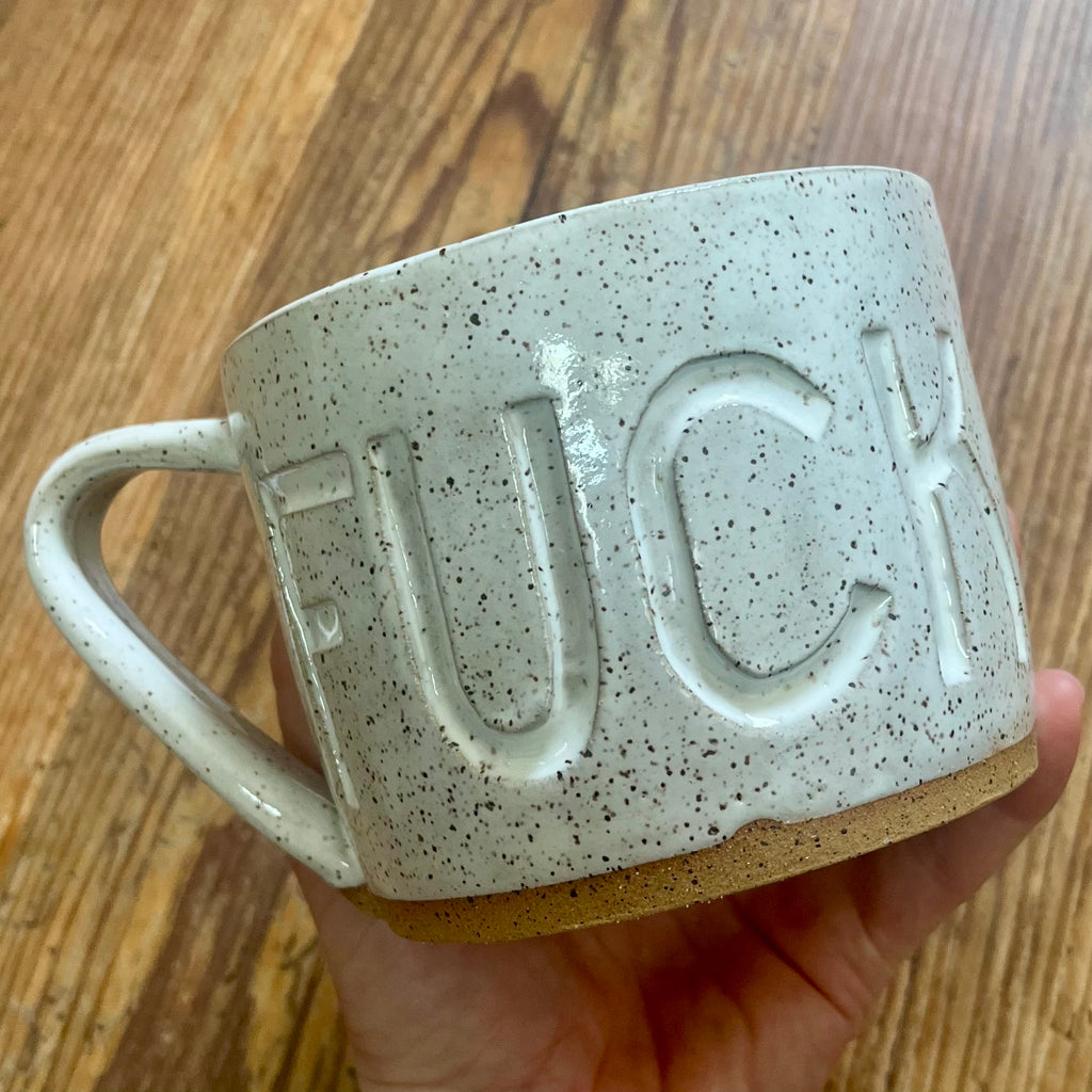 A ceramic mug with a speckled white glaze and carved lettering spelling 'FUCK', held in a person's hand over a wooden table background.