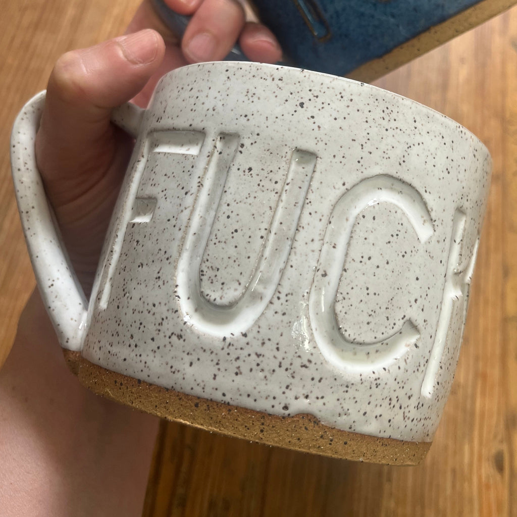 A hand holds two ceramic mugs with the word 'FUCK' embossed on the front, one in speckled off-white and one in glossy blue, over a wooden floor background.