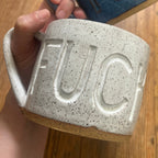 A hand holds two ceramic mugs with the word 'FUCK' embossed on the front, one in speckled off-white and one in glossy blue, over a wooden floor background.